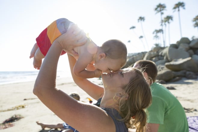 En lykkelig mor løfter opp sin lille baby i luften for et kyss på en solrik strand, med glitrende hav og høye palmer i bakgrunnen.